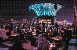 A lively bar scene with people socializing and sitting at tables under a decorative illuminated ceiling, showcasing an urban skyline backdrop.