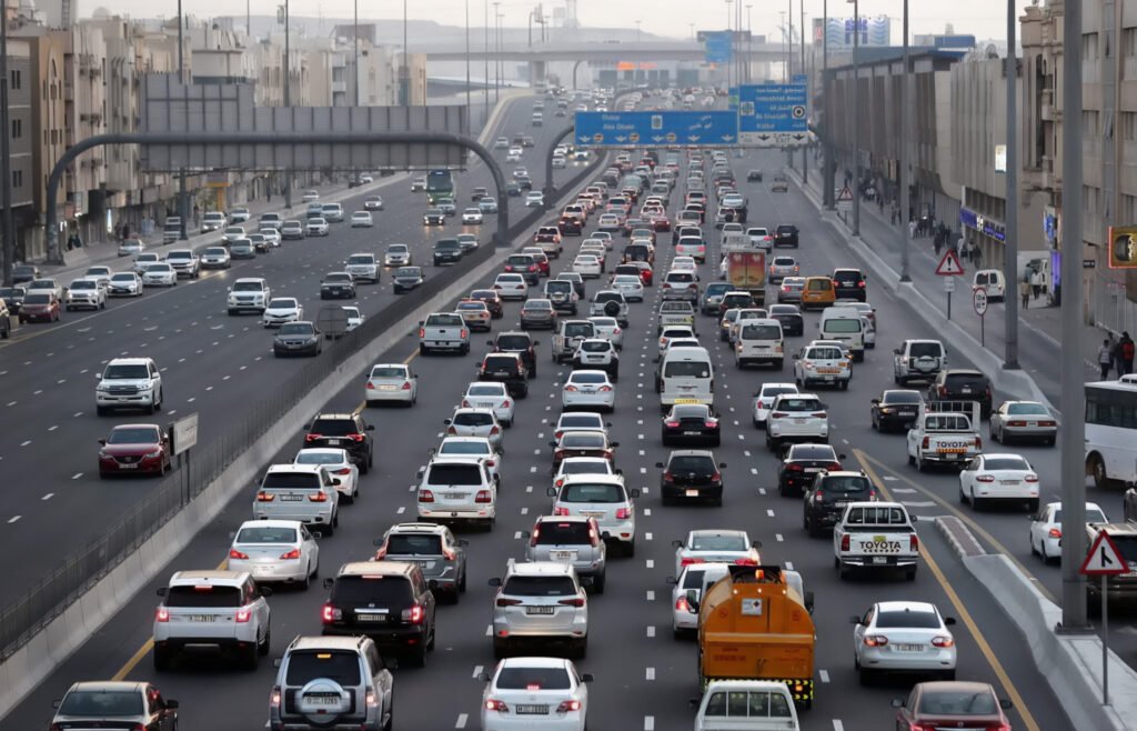 A busy highway with heavy traffic in multiple lanes, showing numerous cars and buses. Overhead signs indicate directions to various locations. Buildings and pedestrian infrastructure line either side of the road.