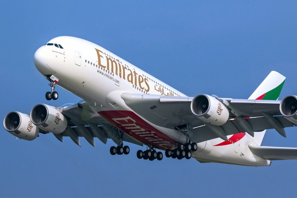 An Emirates Airbus A380 in flight against a clear blue sky, with landing gear extended and the airline's logo visible on the fuselage and engines.