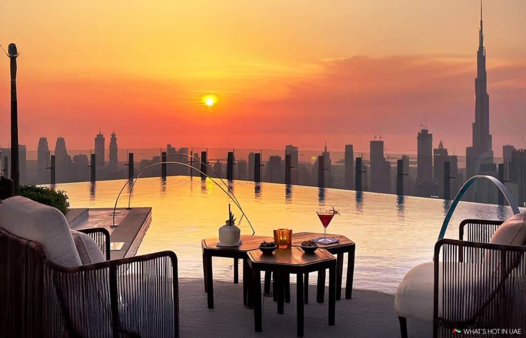 A rooftop infinity pool with a view of a city skyline during sunset, featuring comfortable seating and a table with a drink and snacks in the foreground.