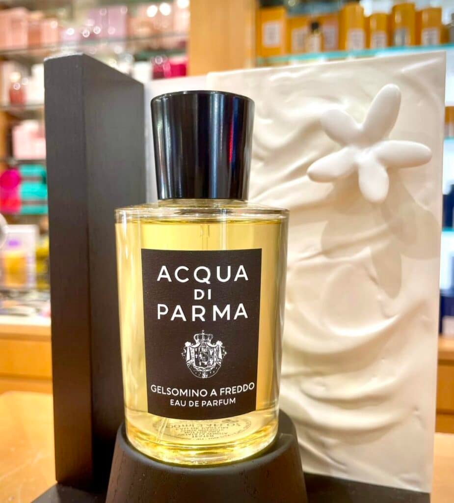 A bottle of Acqua di Parma Gelsomino A Freddo Eau de Parfum displayed on a wooden stand, with a white decorative background featuring a flower design.