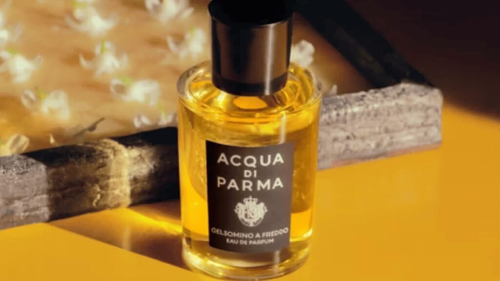 A bottle of Acqua di Parma Gelsomino A Freddo Eau de Parfum with a black cap and label, set on a yellow surface with blurred jasmine flowers in the background.