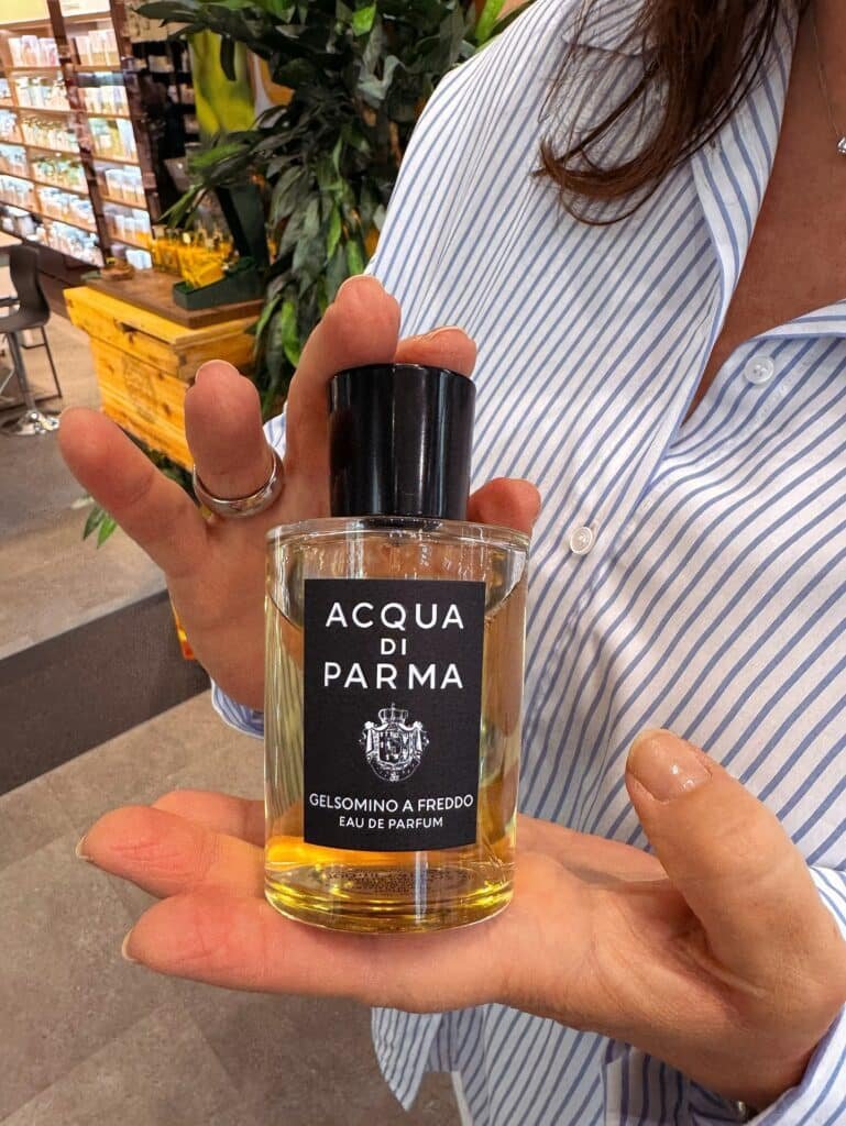 A person holding a bottle of Acqua di Parma Gelsomino a Freddo Eau de Parfum in their hand, with a striped shirt visible in the background.