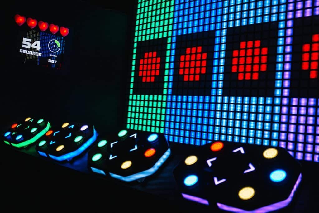 A close-up view of an interactive game setup featuring colorful illuminated buttons and a digital display indicating 54 seconds left and a score of 887. A grid of glowing multicolored squares with red circles is visible in the background.