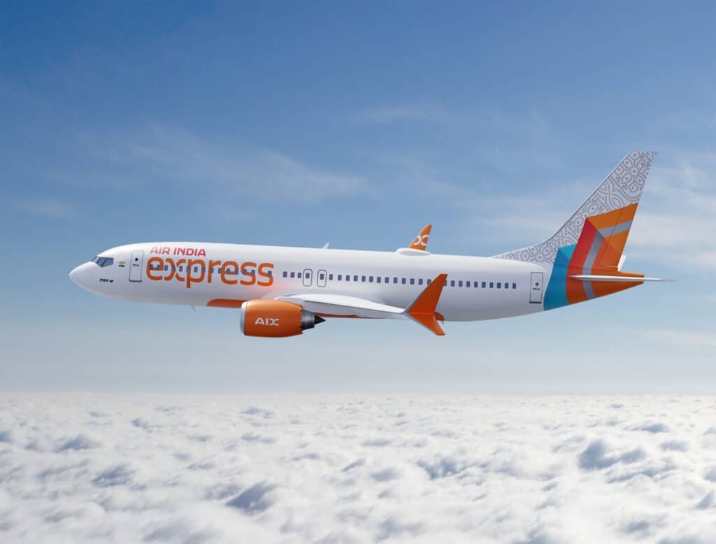 An Air India Express aircraft flying above the clouds against a clear blue sky, featuring orange and white colors with a decorative tail design.