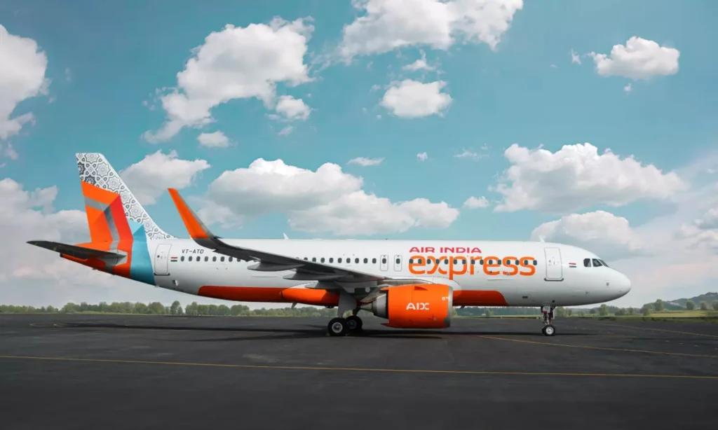 A side view of an Air India Express airplane on the tarmac, featuring orange and white colors with a patterned tail design, under a blue sky with scattered clouds.
