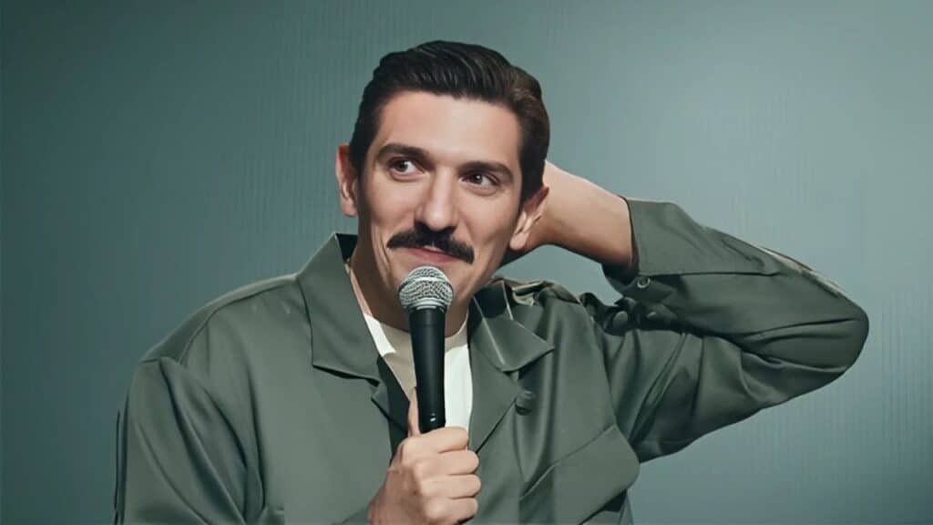 A man with a mustache holding a microphone, wearing a green jacket, with one hand behind his head, against a plain background.