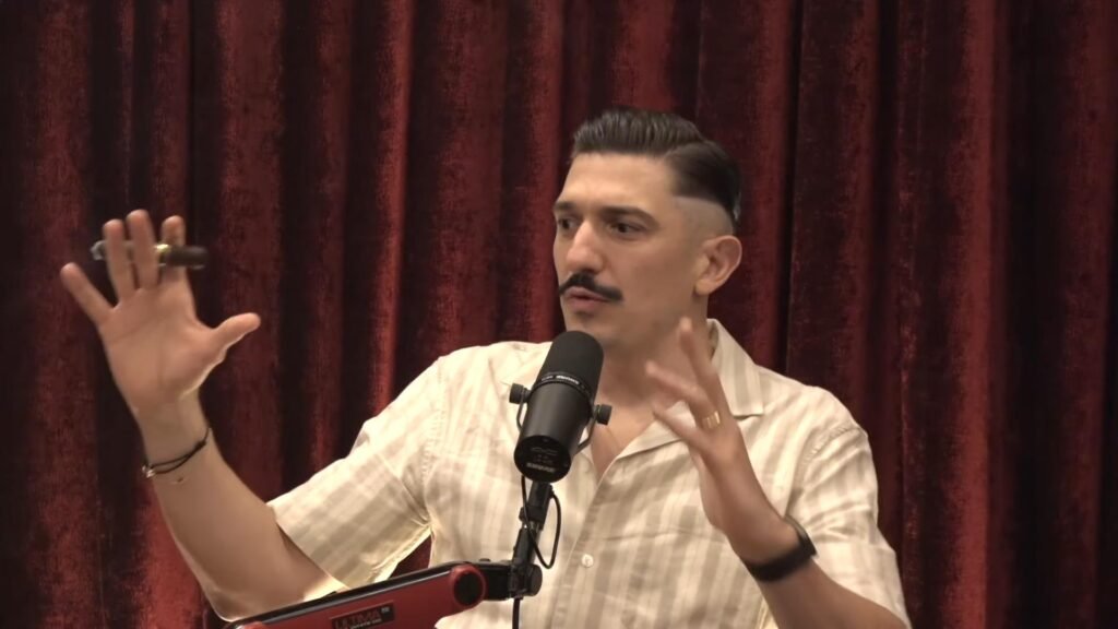 Andrew Schulz is speaking into a microphone, gesturing with his hands, and holding a cigar. He is wearing a light-colored, short-sleeve shirt and is positioned in front of a red curtain background.