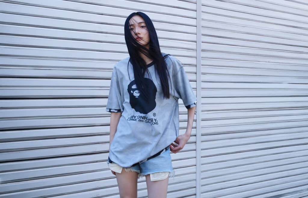 Person wearing a gray oversized BAPE COMME des GARÇONS Osaka T-shirt with a graphic print, standing against a corrugated metal wall with long hair partially covering their face.