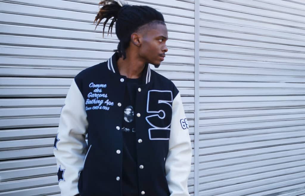 A man wearing a black and white varsity jacket with lettering on the chest and sleeves, standing in front of a metal shutter.