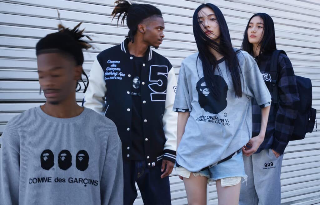 Four people are standing in front of a metal shutter, wearing BAPE clothes featuring "Comme des Garçons" and stylized face designs. The person in the foreground has blurred features and a grey sweater, while others wear a varsity jacket, oversized t-shirts, and casual pants.