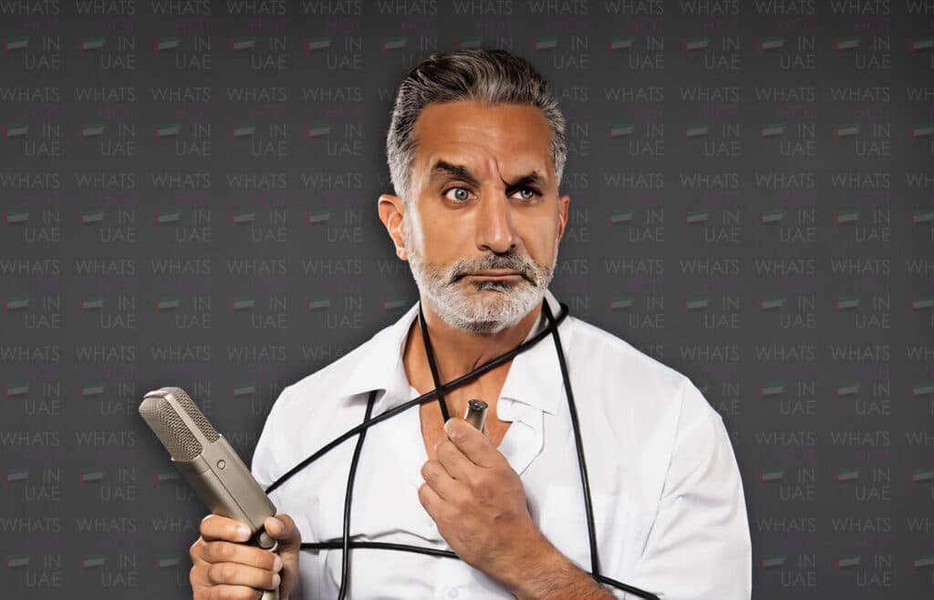 Bassem Youssef stands holding a microphone, with the microphone cable wrapped around his shoulders, against a backdrop that says 'What's Hot in UAE.'