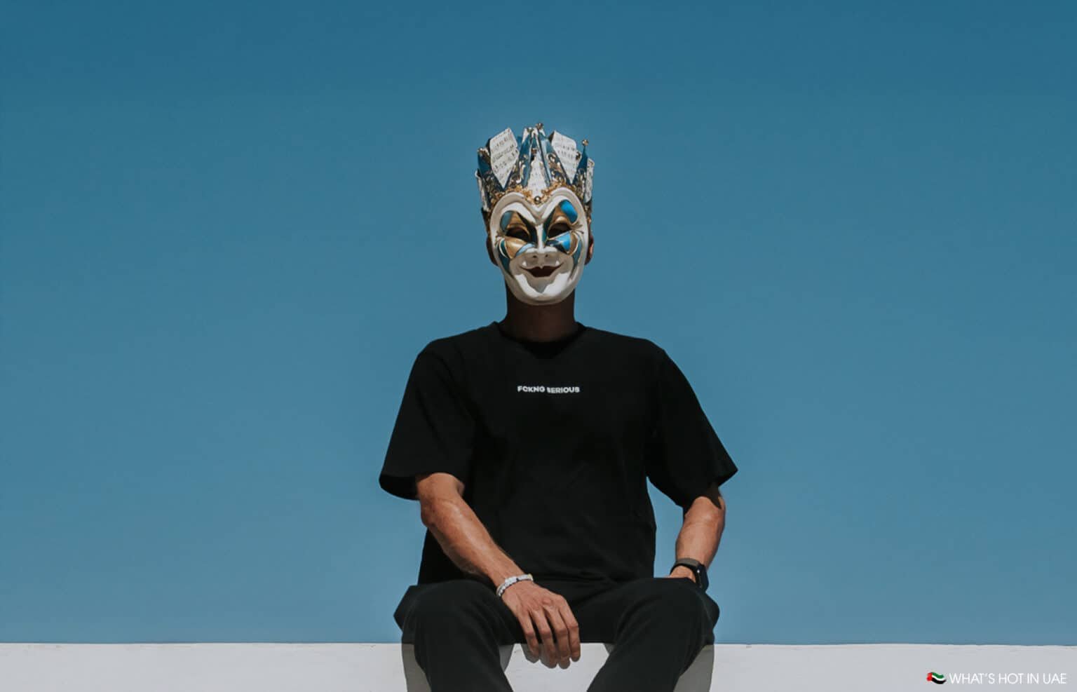 A person wearing a theatrical mask with ornate patterns and a crown is sitting against a clear blue sky, dressed in a black t-shirt and pants.