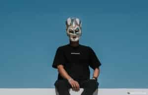 A person wearing a theatrical mask with ornate patterns and a crown is sitting against a clear blue sky, dressed in a black t-shirt and pants.