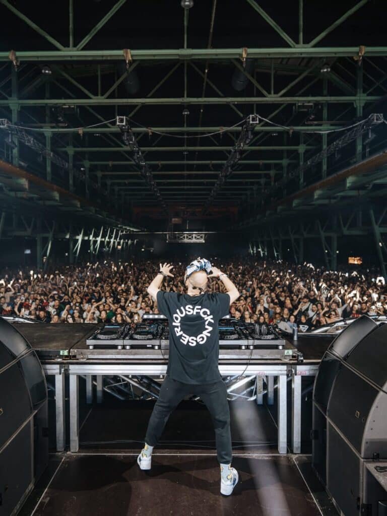 Boris Brejcha on stage facing a large crowd in a packed venue, wearing a black T-shirt and cap, with turntables in front.