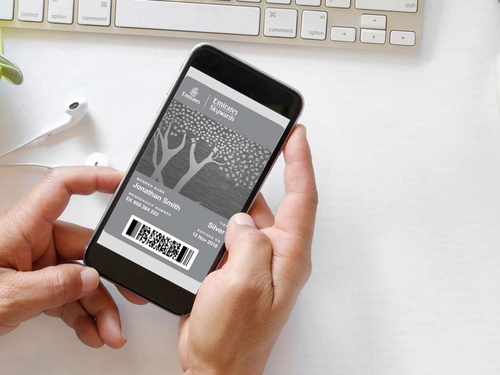 A person is holding a smartphone displaying an Emirates Skywards membership card with membership details and a barcode. The background includes a keyboard and a pair of white earphones on a light-colored surface.