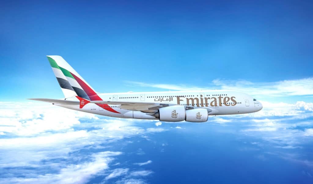 A large Emirates passenger airplane flying above the clouds in a clear blue sky.