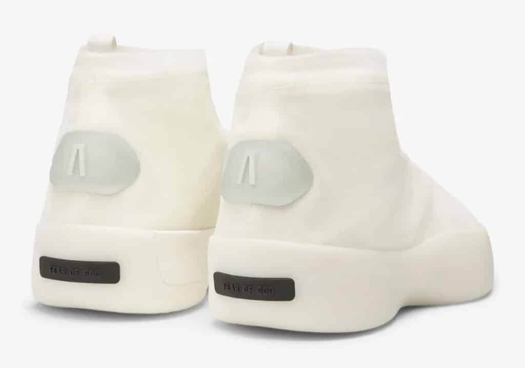 Rear view of white high-top sneakers with 'Fear of God' branding on the heel and a small, semi-translucent patch with a letter design near the ankle.