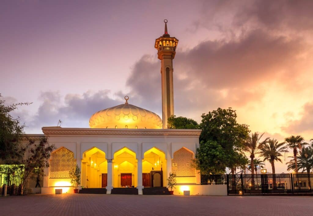 A mosque with a domed roof and tall minaret, illuminated at sunset with palm trees and a dramatic sky in the background.