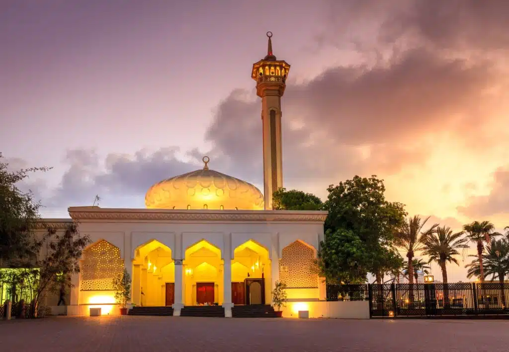 A mosque with a domed roof and tall minaret, illuminated at sunset with palm trees and a dramatic sky in the background.
