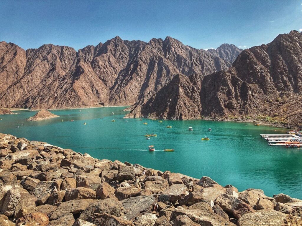 A scenic view of a clear turquoise lake surrounded by rugged, rocky mountains under a clear blue sky, with small boats visible on the water and a rocky shore in the foreground. Hiking Trails UAE Easy Access.