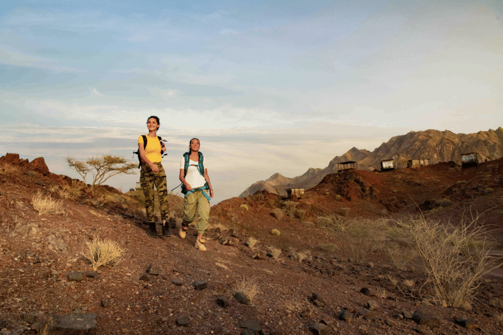 Two women with backpacks hiking in a desert landscape, with rocky terrain and distant mountains under a partly cloudy sky. Hiking Trails UAE Easy Access.