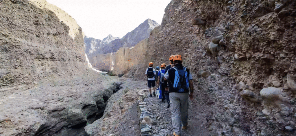 A group of hikers wearing helmets and backpacks walks along a narrow path between steep rock walls in a canyon. Hiking Trails UAE Easy Access.