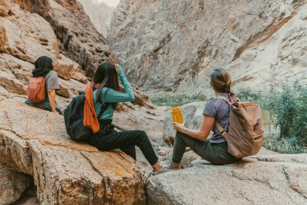 Three people sitting on rocks in a canyon, wearing backpacks and outdoor clothing. One person holds a yellow water bottle. The canyon walls are steep and rocky, with vegetation in the background.