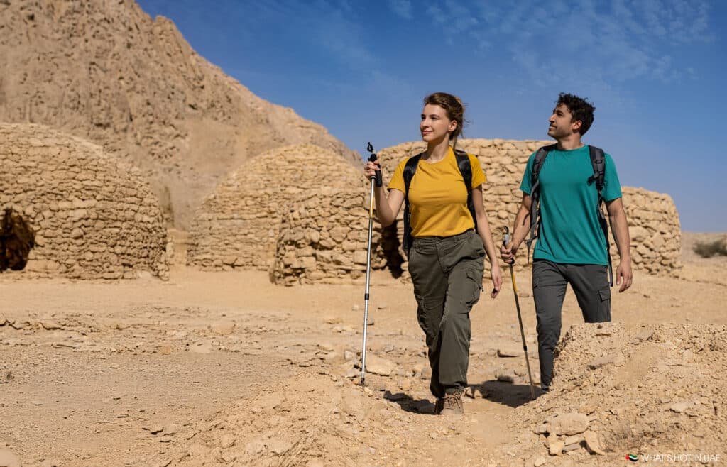Two hikers with backpacks and trekking poles walking through a desert area with stone huts and rocky hills under a clear blue sky.
