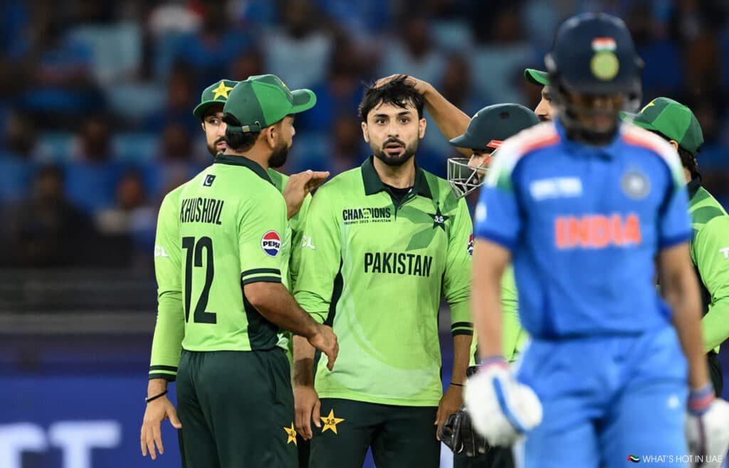 Cricket players from Pakistan's team wearing green uniforms in a group huddle, with a player from India's team wearing a blue uniform walking past in the foreground.