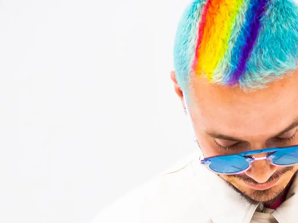 J Balvin in Dubai with rainbow-colored stripes dyed into their light blue hair, wearing blue sunglasses and looking down.