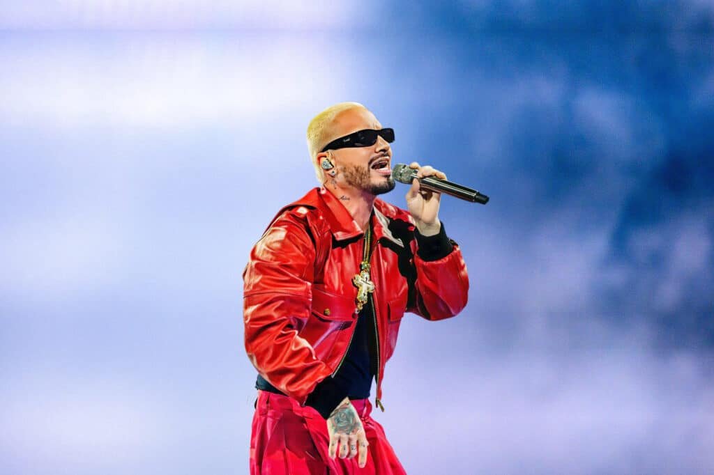 J Balvin singing on stage, wearing a bright red leather jacket, matching red pants, and dark sunglasses, holding a microphone against a blue background.