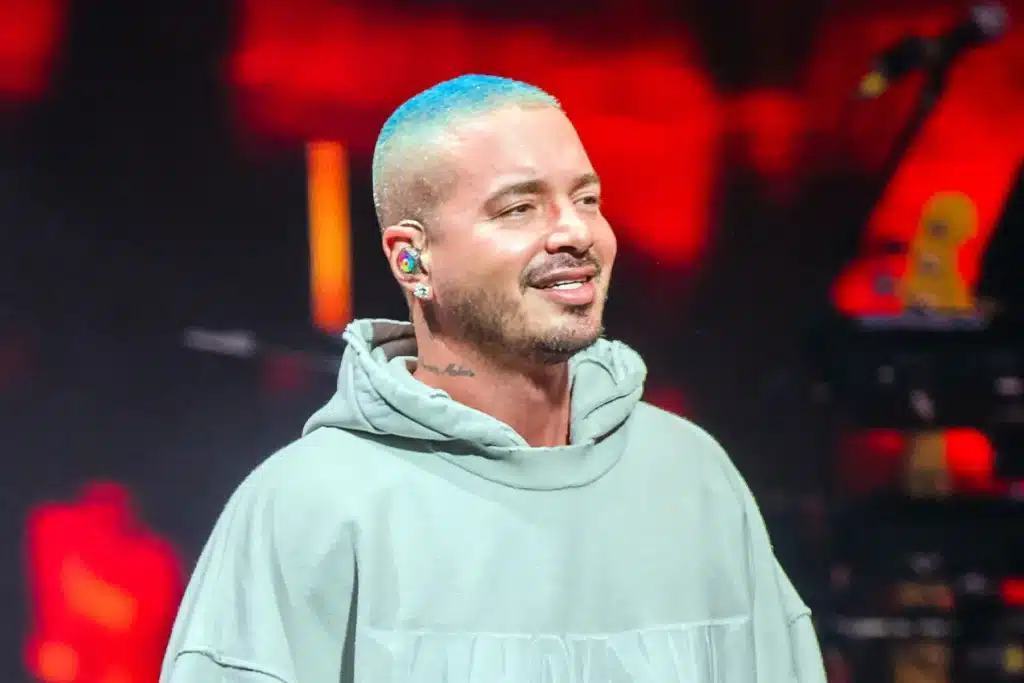 A person with short blue hair wearing a light gray hoodie and colorful earphones, standing against a blurred dark background with red lighting.