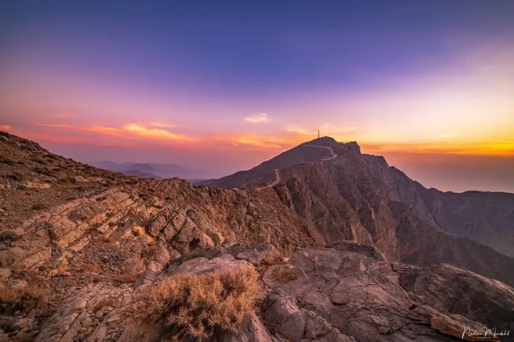 A mountainous landscape at sunset, featuring rugged rocky terrain in the foreground and a peak with a winding road in the distance under a vibrant sky.