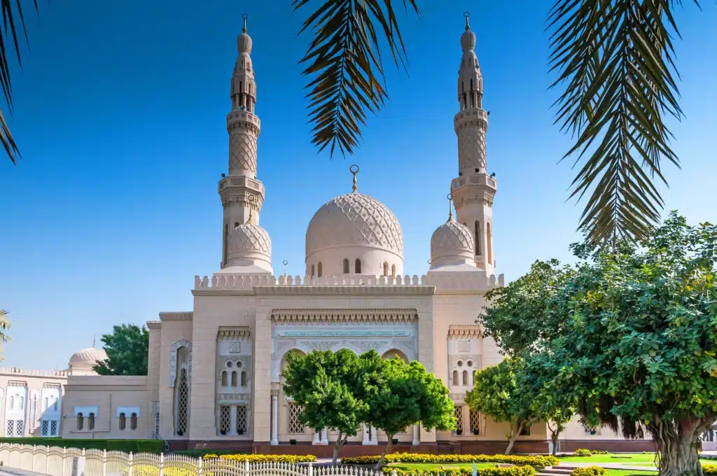 A large mosque with two ornate minarets and a central dome, surrounded by trees and palm fronds against a clear blue sky. Prayer Time in Dubai