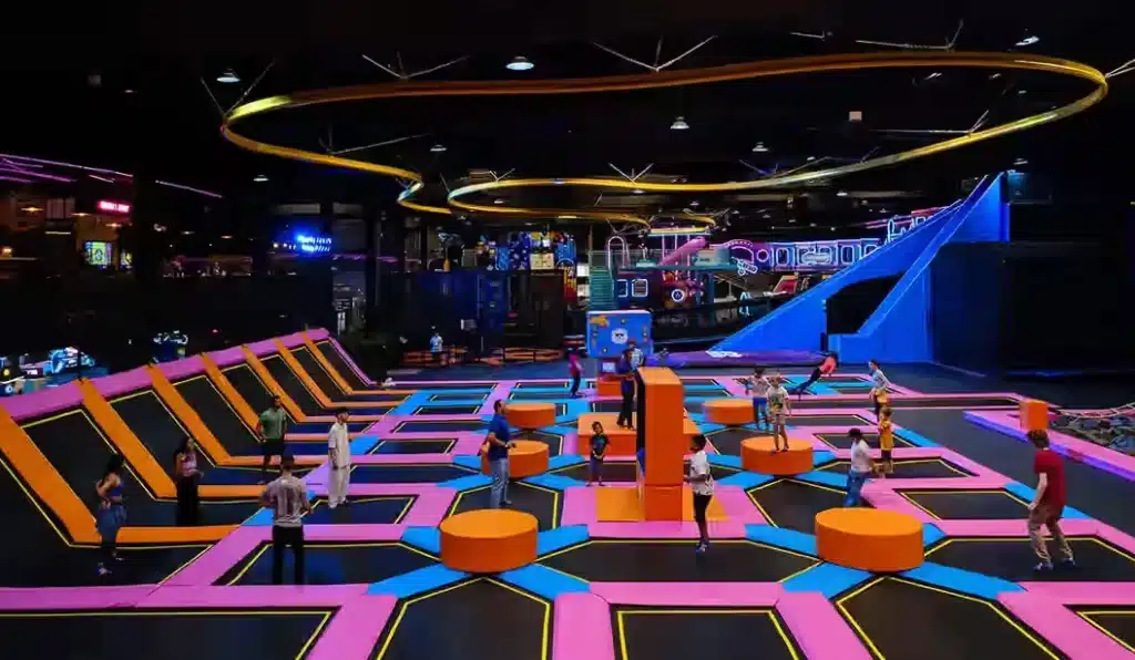 A vibrant indoor trampoline park with colorful pink and orange designs, featuring multiple trampolines and platforms. Various people, including children and adults, are actively playing and jumping. In the background, there is a large blue slide and other play structures.