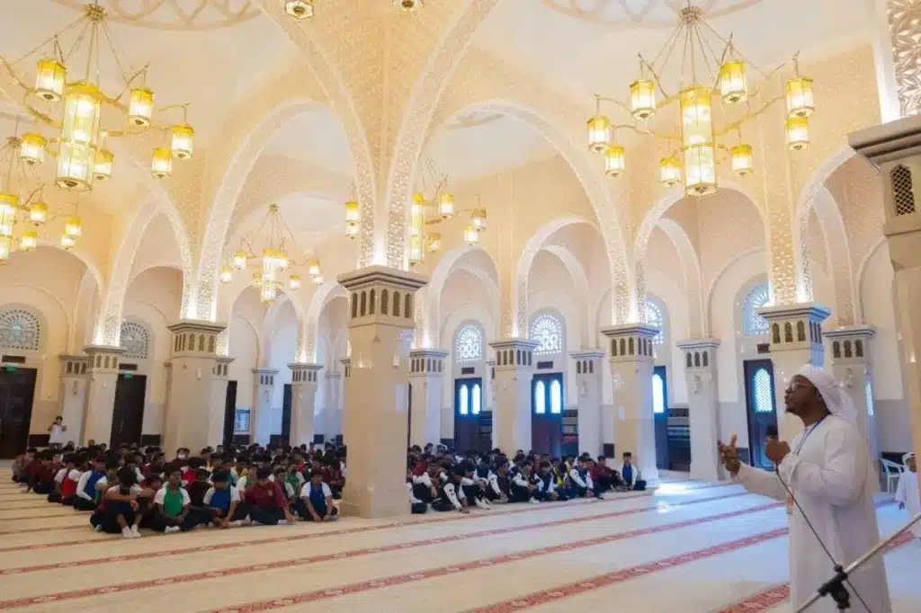 A large, ornate mosque interior with multiple arched columns and chandeliers, where a group of students sit on the carpeted floor, listening to a speaker dressed in traditional white attire. Prayer Time in Dubai