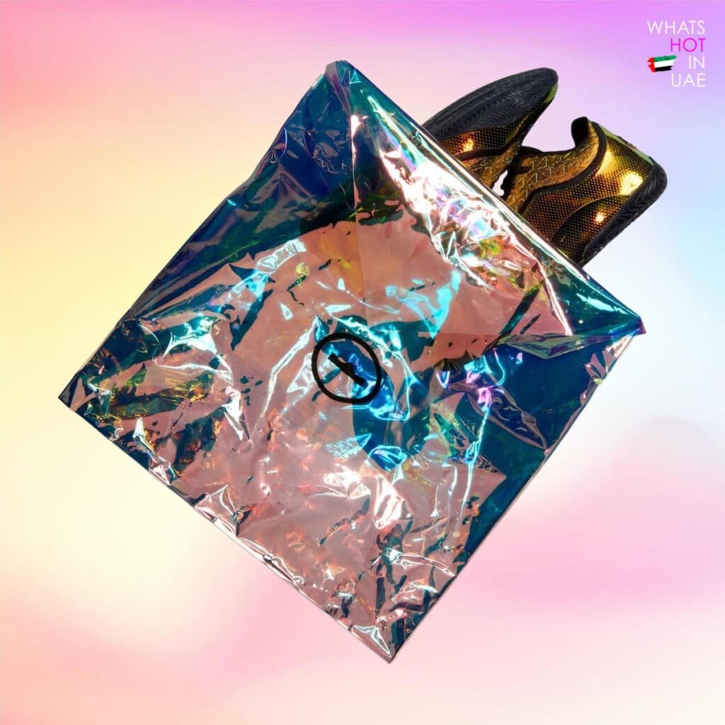 Pair of black and gold sneakers partially inside a reflective, iridescent bag on a gradient pink and yellow background, with text "WHAT'S HOT IN UAE" in the corner.