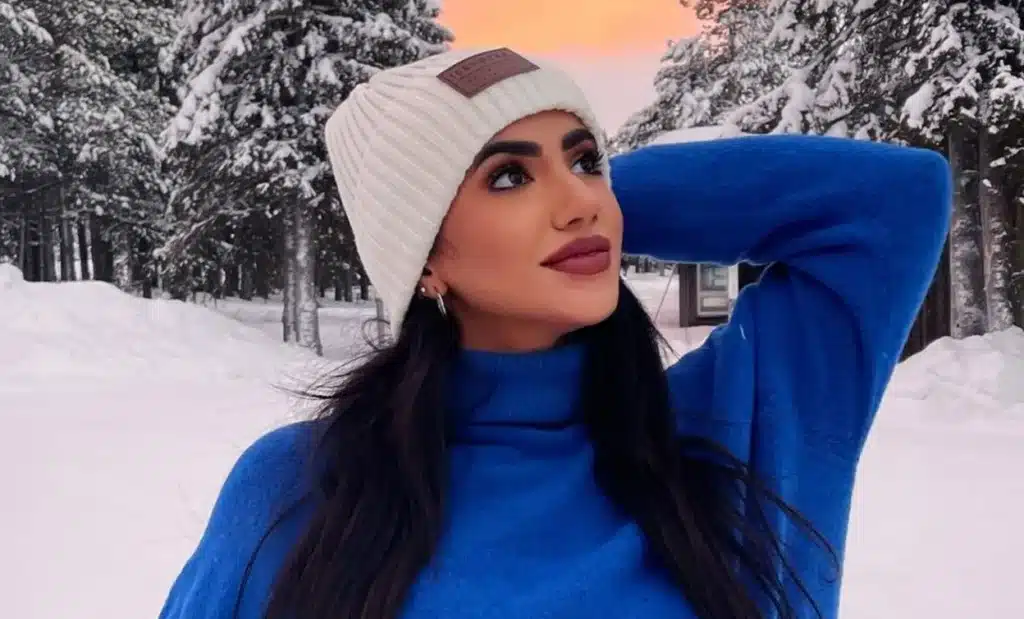 Person wearing a white beanie and blue sweater, standing in a snowy forest with snow-covered trees in the background and an orange-hued sky. Top 25 MENA Social Media Influencers. 