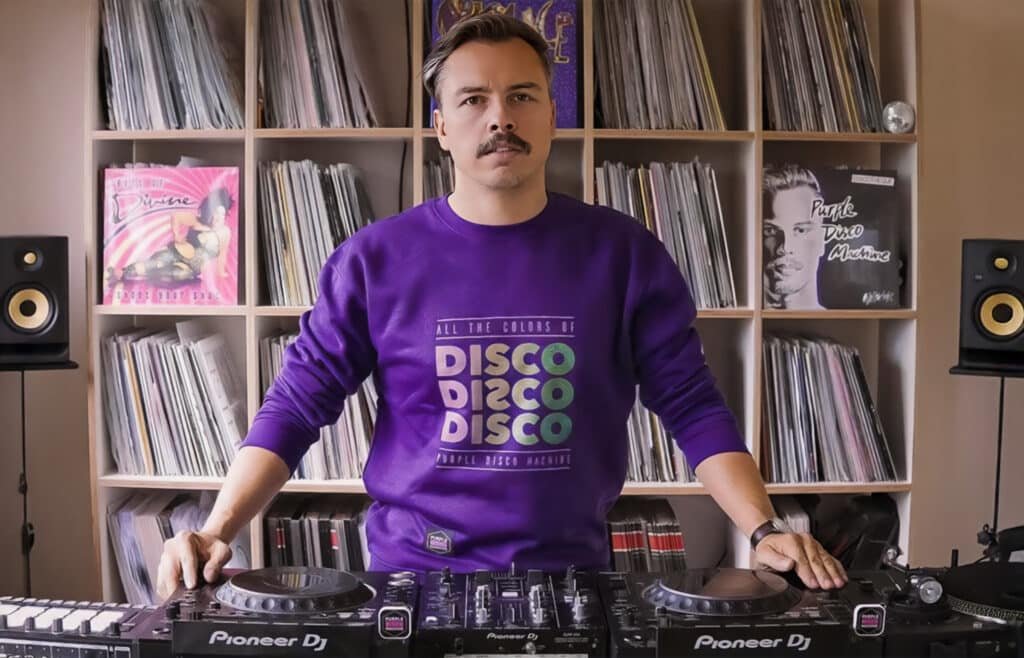 A person wearing a purple sweatshirt with colorful text reading 'DISCO' stands in front of a DJ setup surrounded by shelves filled with vinyl records and speakers.