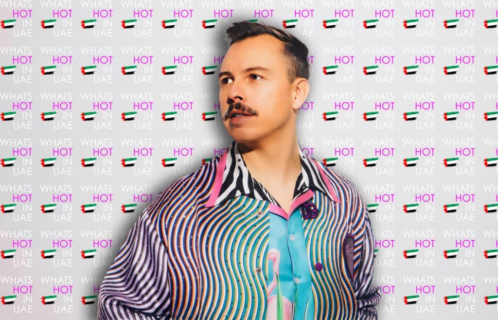 A person wearing a colorful, patterned shirt stands in front of a backdrop featuring the text 'WHAT'S HOT IN UAE' repeatedly, along with small UAE flags.