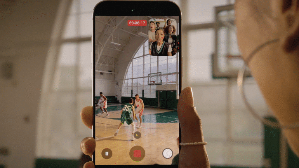 A close-up view of a smartphone screen displaying a live video recording of a basketball game. The phone shows two players in red and white uniforms defending a player in a green uniform with the number 35. A video call is in progress, with a small window showing three people watching the game. The gymnasium is visible in the background through the phone screen. iPhone 17
