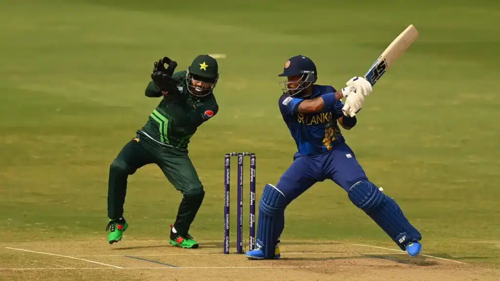 A cricket match in progress with a Sri Lankan batsman in blue and yellow gear playing a shot, while a Pakistani wicketkeeper in green and black uniform is poised to catch the ball. The scene is set on a cricket field with stumps visible in the background.