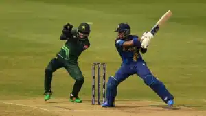 A cricket match in progress with a Sri Lankan batsman in blue and yellow gear playing a shot, while a Pakistani wicketkeeper in green and black uniform is poised to catch the ball. The scene is set on a cricket field with stumps visible in the background.