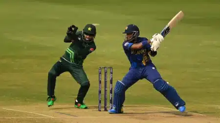 A cricket match in progress with a Sri Lankan batsman in blue and yellow gear playing a shot, while a Pakistani wicketkeeper in green and black uniform is poised to catch the ball. The scene is set on a cricket field with stumps visible in the background.