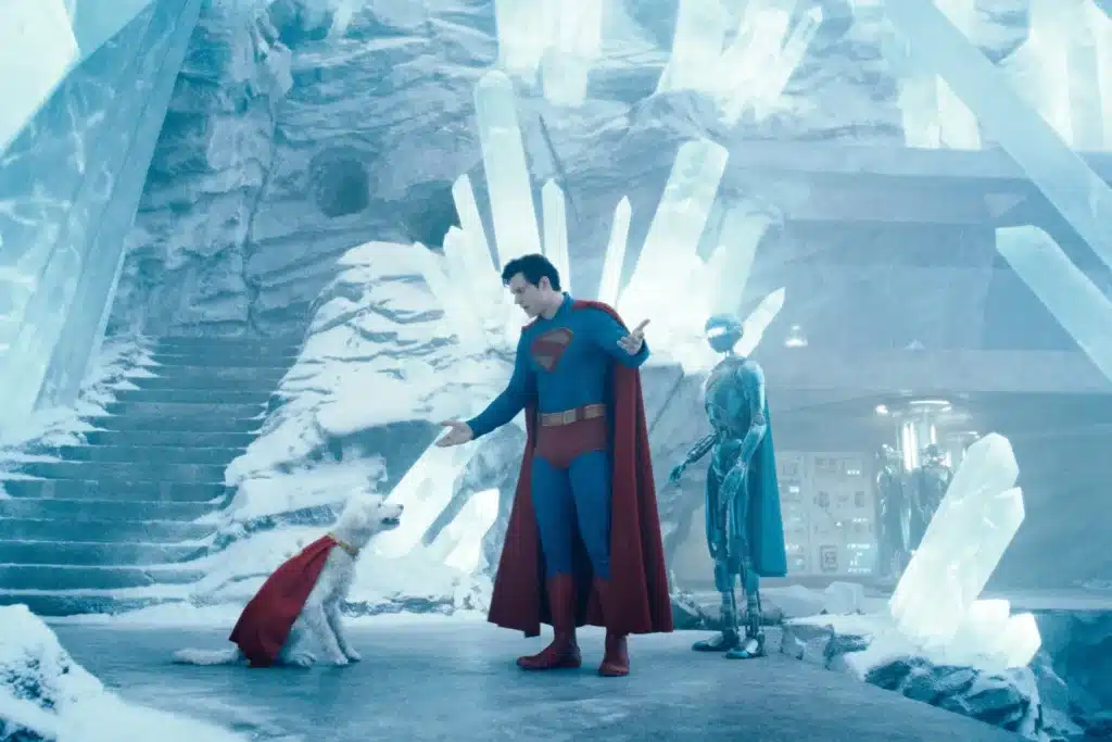 Superman stands inside his crystalline fortress, interacting with a white dog wearing a red cape. A humanoid robot is standing nearby, and the surroundings are icy with large crystal formations.
