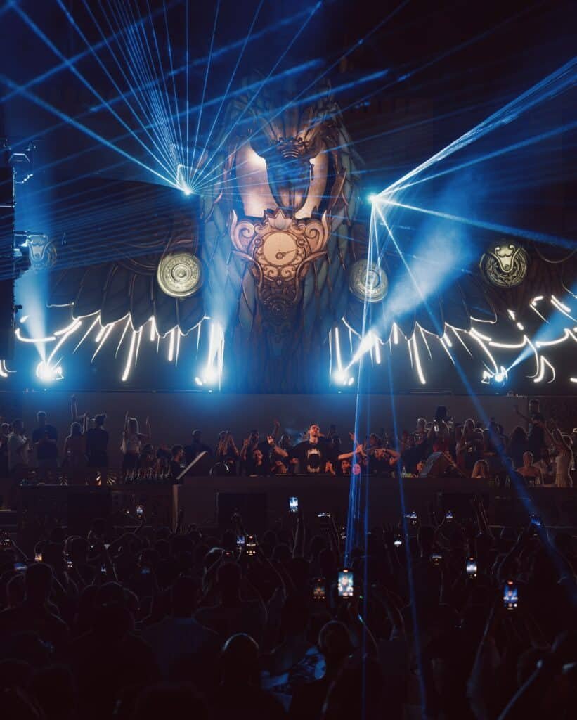 The Terra Solis stage with an elaborate backdrop of decorative owl-like features and blue laser lights emanating in various directions, while a DJ performs for a large crowd of people holding up phones and dancing.