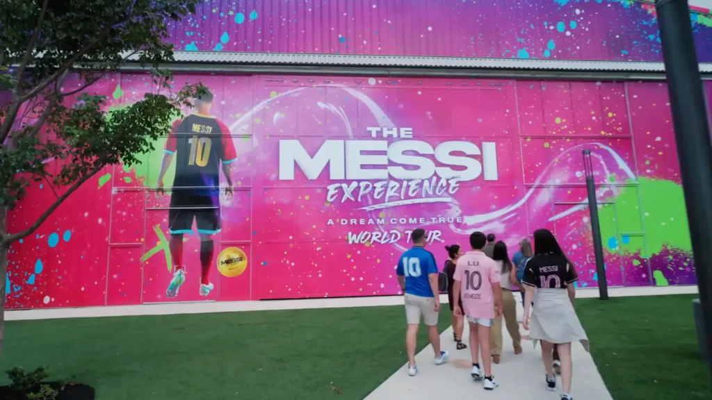 A group of people walking towards a large, colorful advertisement with the text 'The Messi Experience: A Dream Come True World Tour.' The ad features an image of a player wearing a jersey with the name 'Messi' and the number 10. Family and Kids Activities in Dubai