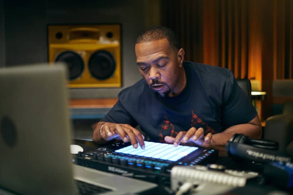 Timbaland in a studio setting concentrates while using a music production device with illuminated pads, surrounded by studio equipment and a pair of headphones.