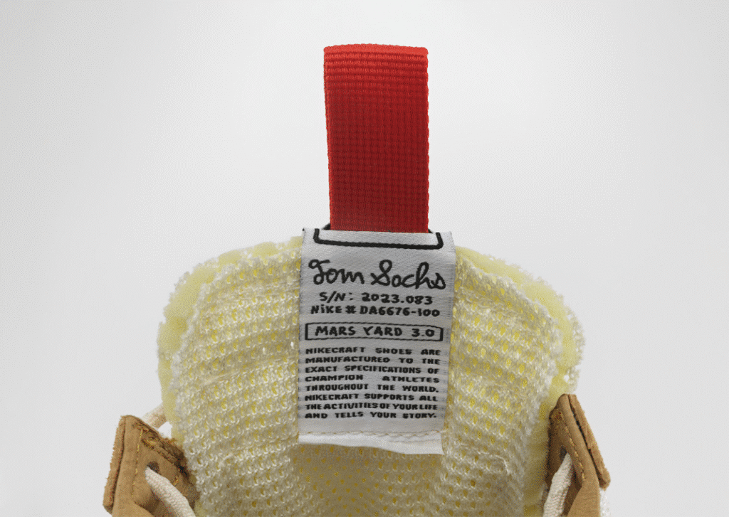 Close-up of a sneaker's label with a red pull tab, showing the text "Tom Sachs, S/N: 2023.083, Nike DA6676-100, Mars Yard 3.0" and additional details printed below. Tom Sachs Mars Yard 3.0